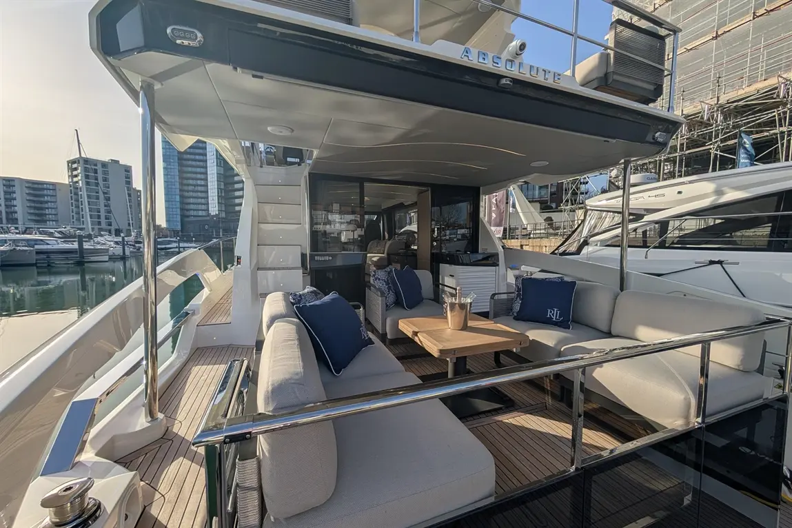 Spacious yacht deck with grey seating, wooden table, and glass railings, overlooking a marina with modern buildings and boats.